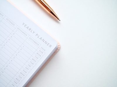 The corner of a book- with the words, "Yearly Planner" at the top of the open page- is visible from the left side of the frame. The book sits on a white background. Above the book is a golden pen.