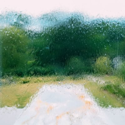 A path leading to a gathering of trees who leaves are bright and green. The scene is obscured by a layer of rain as if the viewer is looking out a window