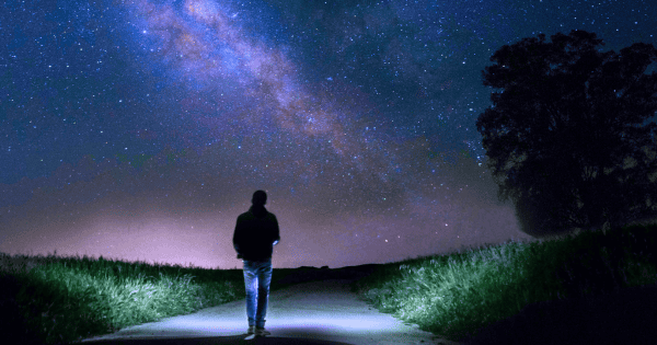 silhouette of a singular man in jeans walking down the middle of a road with the milky way and stars above him