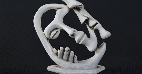 An abstract sculpture in white of portions of two faces in a romantic pose. A frontal view of the closed right eye, nose, lips, chin, and left cheek surrounded by a swoop suggesting the shape of hair is being embraced by a face in profile whose hand can be seen wrapping around the missing right cheek. A kiss seems imminent.