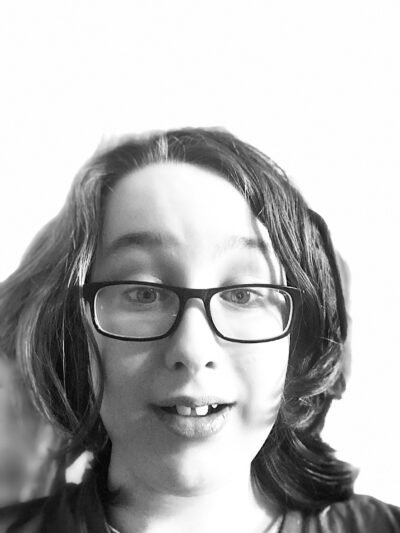 A black and white image of a boy with shaggy hair and glasses. His eye-brows are arched and he's gazing intently at the camera and half-smiling.