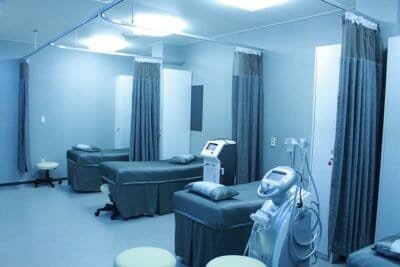 A sterilized, pearl-white hospital room with three gray beds and open divider curtains lining the wall