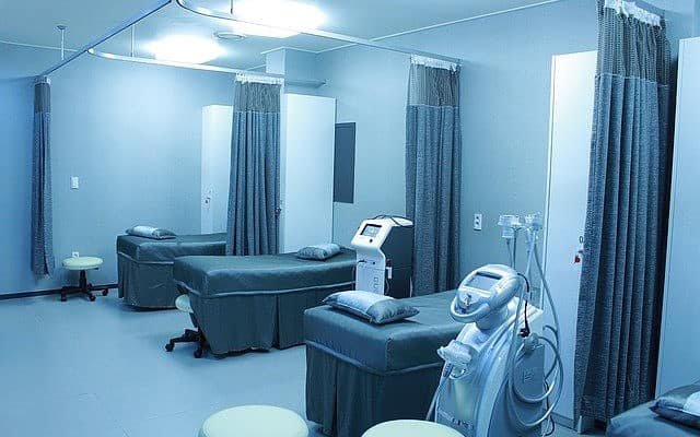 A sterilized, pearl-white hospital room with three gray beds and open divider curtains lining the wall