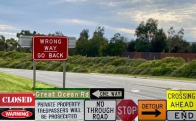 The image is of an empty road, and road sign saying, 'Wrong Way, Go Back", and along the lower part of the image, there numerous other roads signs