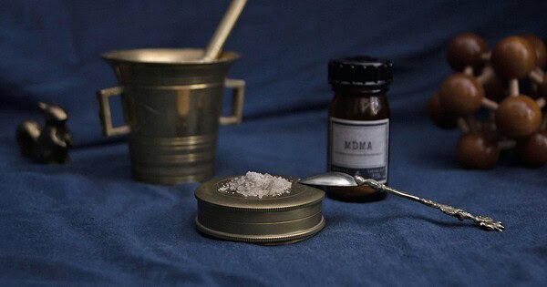A white crystal powder sits on a brass container. A spoon leans against the container. The lab chemical bottle behind the container, powder, and spoon says "MDMA". To the left of these items is a brass mortar and pestle. To the far left in background is a brass squirrel. To the far right in the background is a wooden dodecahedron made out of balls and rods. All of these items sit on a blue table cloth in front of a blue backdrop.