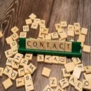 A brown table with light brown tiles with letters scattered around a green board holding tan block letters that spell out the word contact.