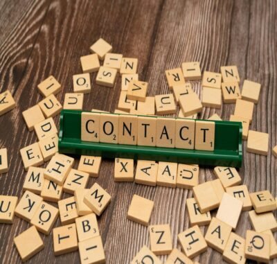 A brown table with light brown tiles with letters scattered around a green board holding tan block letters that spell out the word contact.