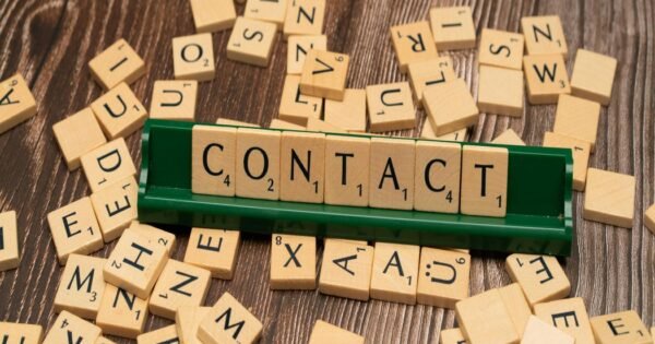 A brown table with light brown tiles with letters scattered around a green board holding tan block letters that spell out the word contact.