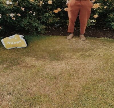 A man standing in front of a bush wearing brown shoes in grass. A small yellow material next to him that reads happy.