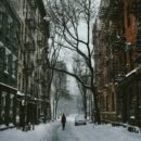 A street blanketed with snow between two buildings and one tree. A single bundled up person is shown.