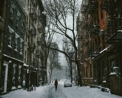 A street blanketed with snow between two buildings and one tree. A single bundled up person is shown.