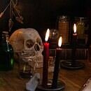 A skull sits on a table surrounded by glass potion bottles and red candles lit with an eerie glow