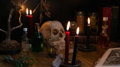 A skull sits on a table surrounded by glass potion bottles and red candles lit with an eerie glow