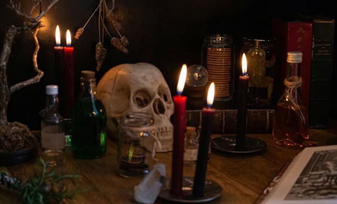 A skull sits on a table surrounded by glass potion bottles and red candles lit with an eerie glow