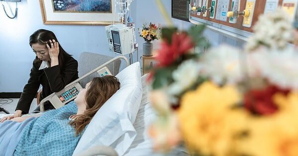 Two people in hospital room. One in bed. The other sitting next to the bed. Flowers out of focus in front.