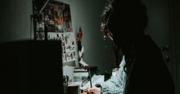 A woman in a gray knit sweater sitting with dim lighting at their desk writing in a notebook.