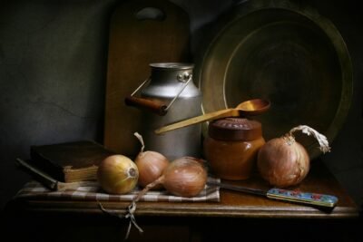 Four fresh onions on a vintage kitchen table with a milk can, jar, spoon, knife, and cookbooks