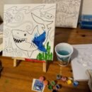 Canvas with shark partly painted. Paint supplies on table.