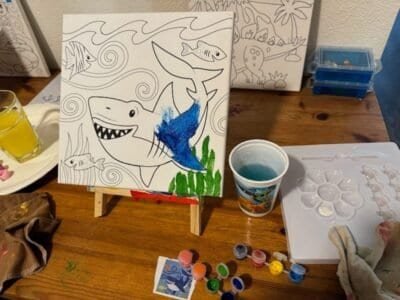 Canvas with shark partly painted. Paint supplies on table.