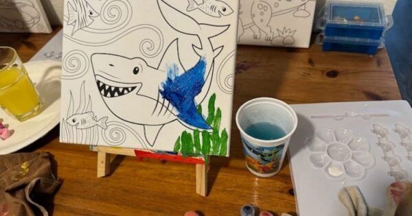 Canvas with shark partly painted. Paint supplies on table.