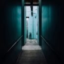 A hallway with green walls and gray floors that shows a lit baby blue brick wall and window in the distance.