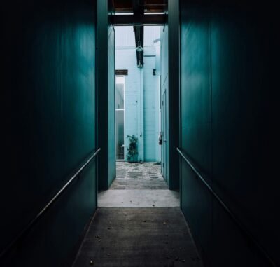 A hallway with green walls and gray floors that shows a lit baby blue brick wall and window in the distance.