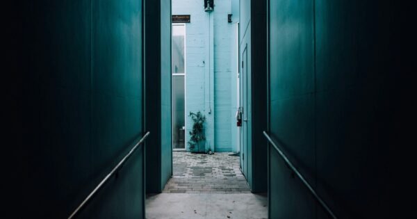 A hallway with green walls and gray floors that shows a lit baby blue brick wall and window in the distance.