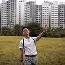 Man in white shirt holding up arm to stop. Standing on grass field in front of tall buildings.