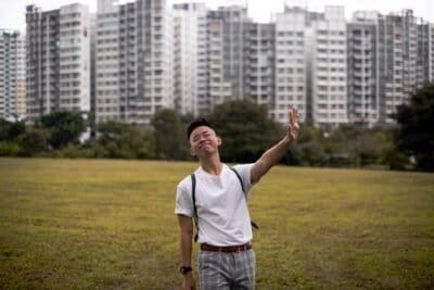 Man in white shirt holding up arm to stop. Standing on grass field in front of tall buildings.