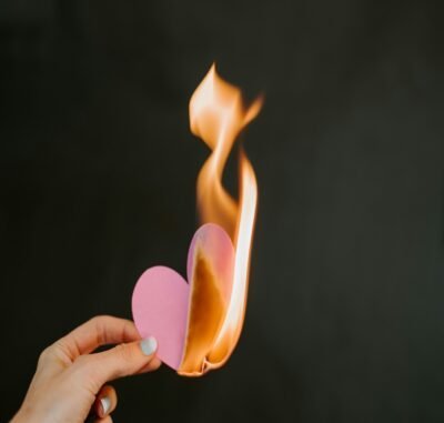 A hand holding a pink paper heart on fire with a bright orange flame.