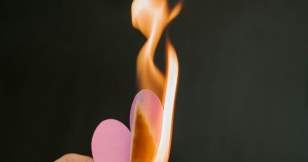 A hand holding a pink paper heart on fire with a bright orange flame.