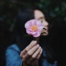 A dark haired woman holding up a pink flower with her eyes closed.