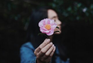 A dark haired woman holding up a pink flower with her eyes closed.