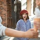 Girl in red hat looking excitedly at two arms holding ice cream.