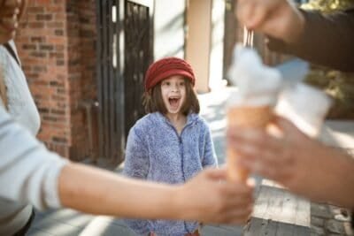 Girl in red hat looking excitedly at two arms holding ice cream.