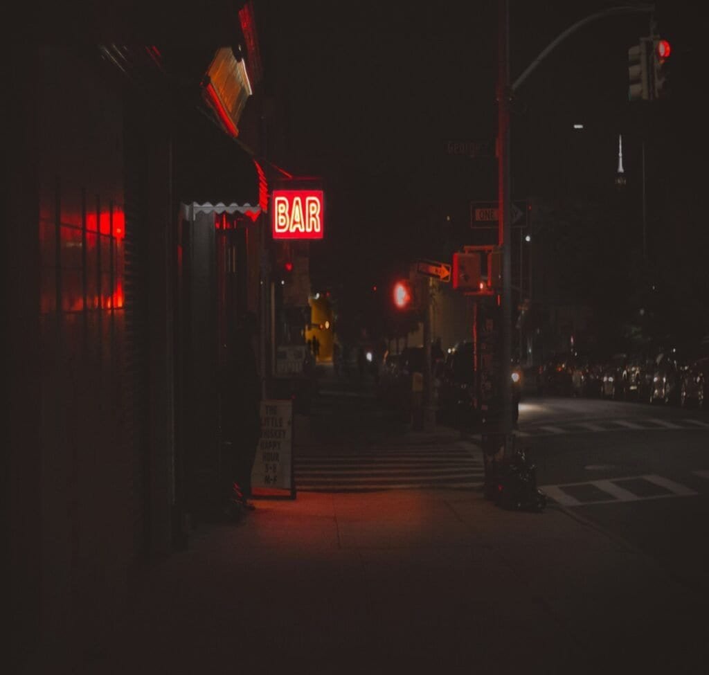 A dark street with cars and a building to the left with a neon sign that reads bar.