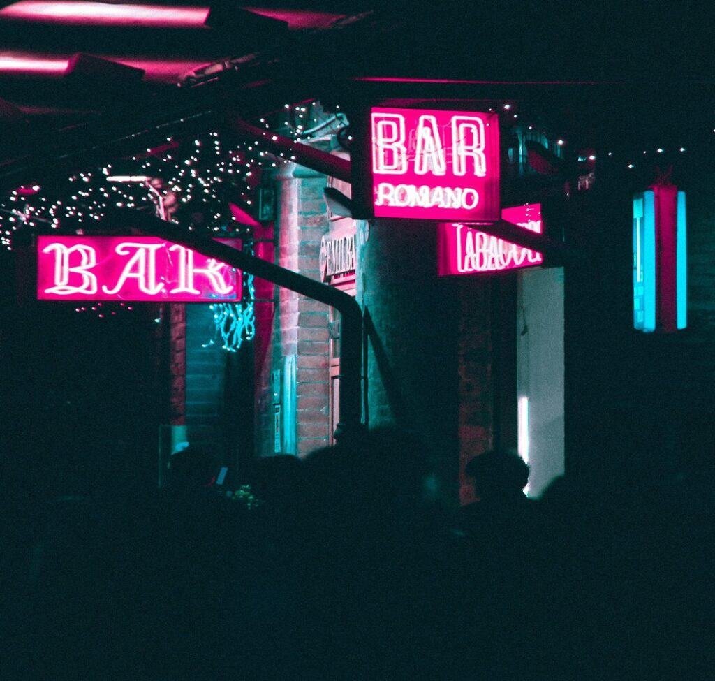 A dark bar with shadows of people and two pink neon signs that read bar.