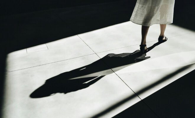 Woman in a white dress walking in a dark lit area with her shadow behind her.
