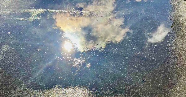 The image is of a shimmering puddle on a concrete path, which is mirroring the sky above