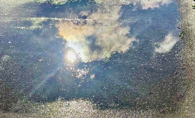 The image is of a shimmering puddle on a concrete path, which is mirroring the sky above