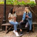Two women sitting on a brown wooden bench holding sodas