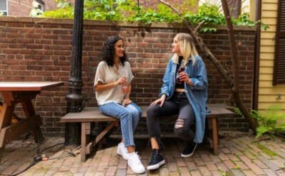 Two women sitting on a brown wooden bench holding sodas