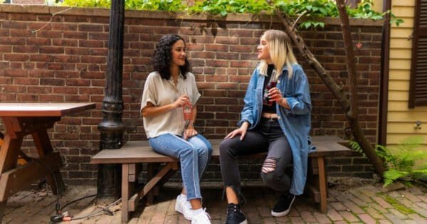 Two women sitting on a brown wooden bench holding sodas