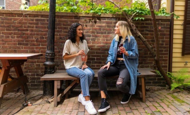 Two women sitting on a brown wooden bench holding sodas