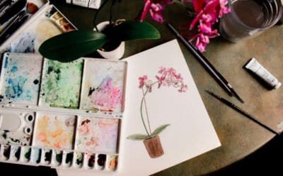 White, pink, and green watercolor floral painting on a table with paintbrushes and paint.