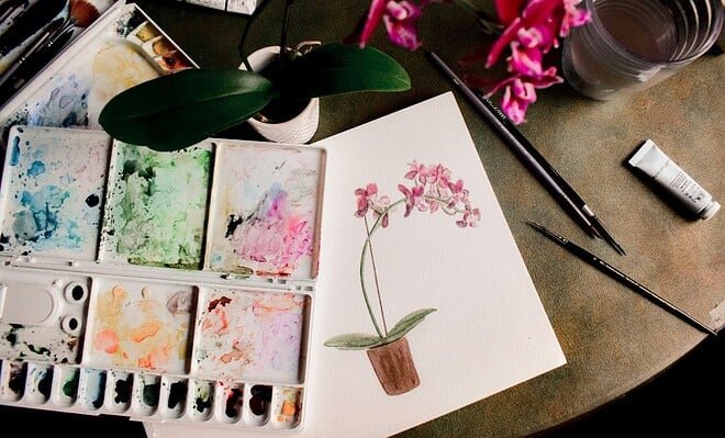 White, pink, and green watercolor floral painting on a table with paintbrushes and paint.