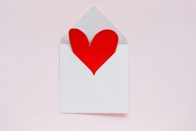 There is a pink background and a small white envelope that is opened, a red paper heart sticking out of it