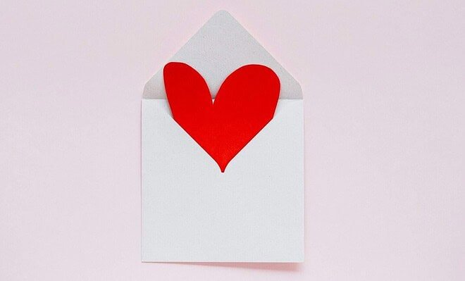 There is a pink background and a small white envelope that is opened, a red paper heart sticking out of it