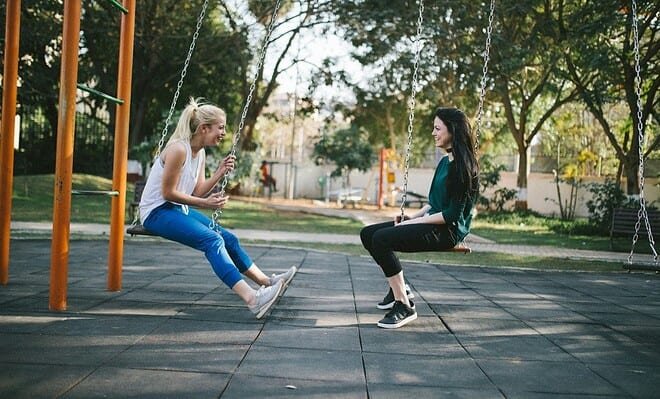 Two women at a park sitting on swings and smiling while having a conversation