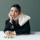 Asian woman with scarf wrapped around her neck sitting, elbow on table with her hand resting on her cheek.